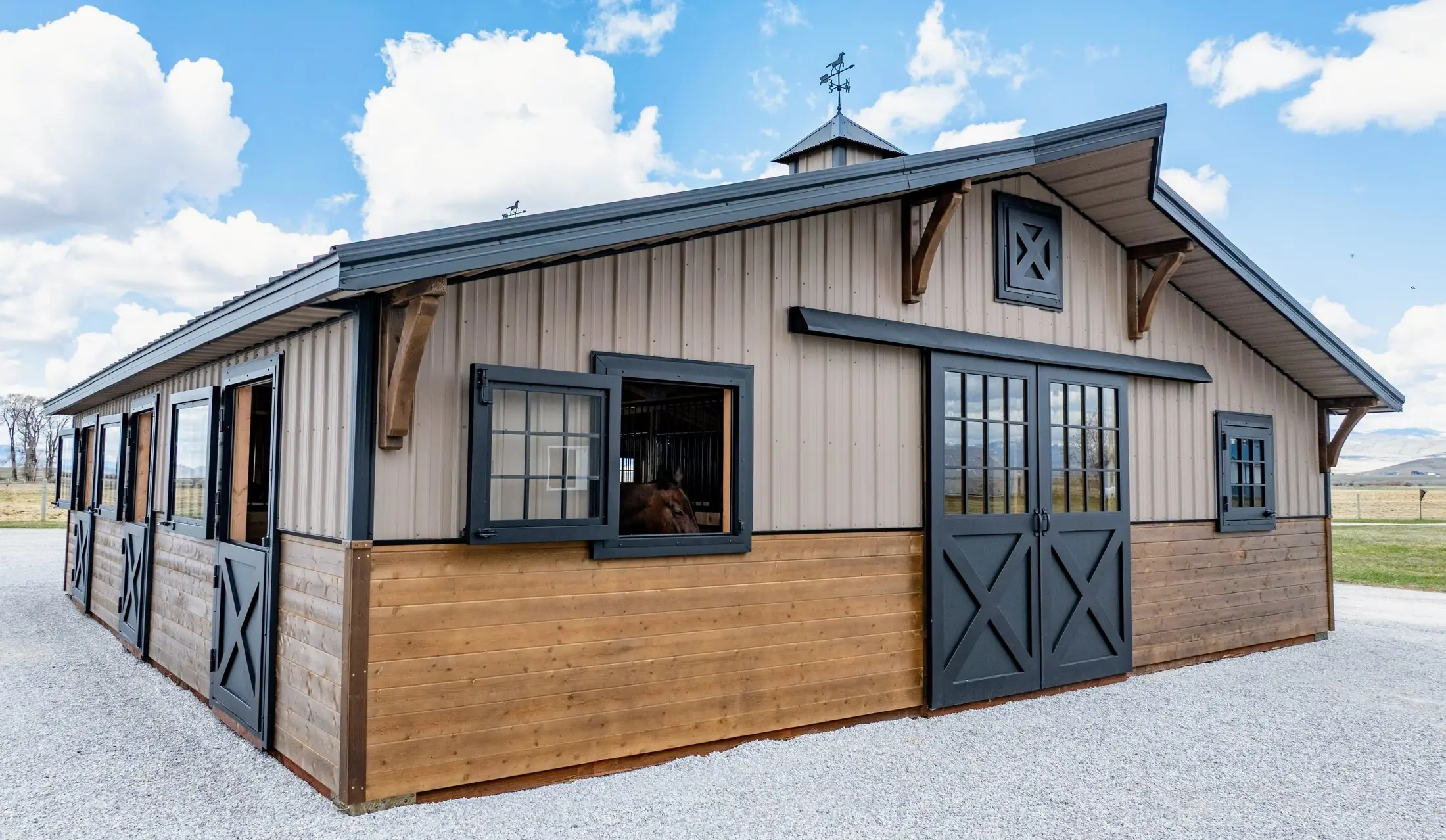 34×36 two-piece horse barn, wide three-quarter exterior view, a horse at the stall window, mountains behind